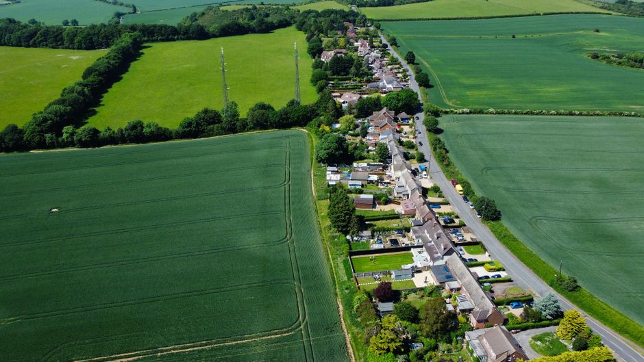 An aerial view of a small village with a row of terraced houses and gardens along a narrow main road, surrounded by expansive green fields and farmland. The gardens feature well-maintained lawns, flow